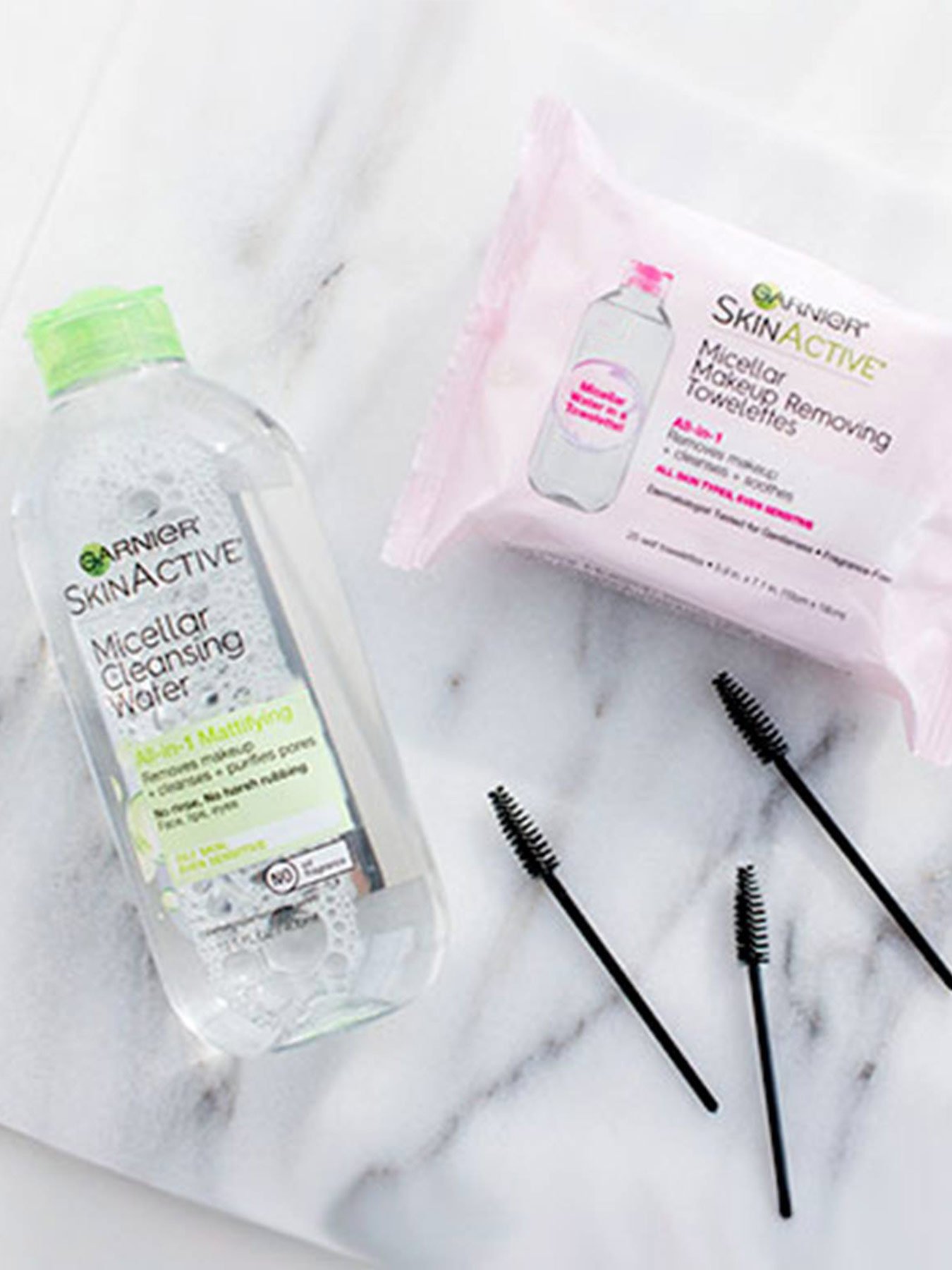 Garnier SkinActive Micellar Cleansing Water All-in-1 Mattifying and SkinActive Micellar Makeup Removing Towelettes on a white marble board with three eyelash brushes on a white surface.